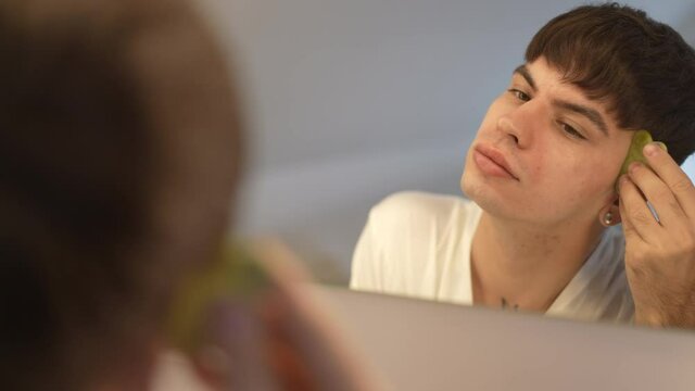 Reflection in mirror of confident LGBT man massaging face with gua sha in slow motion. Portrait of young handsome non-binary person taking care of facial skin at home indoors in the morning