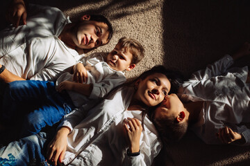 Happy family dressed in jeans and white shirts lie on the floor together in sunlight. 