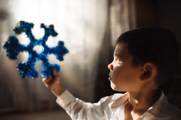 A boy in a white shirt looks at a Christmas tree decoration in the form of a blue snowflake