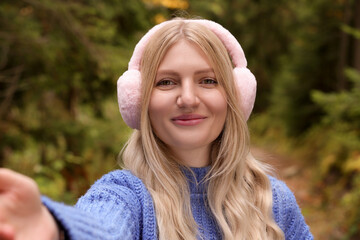Young woman in warm earmuffs taking selfie outdoors on autumn day