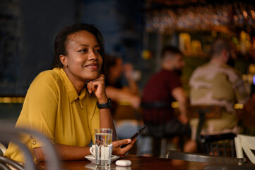 African american woman using a smartphone and drinking coffee while sitting in a cafe