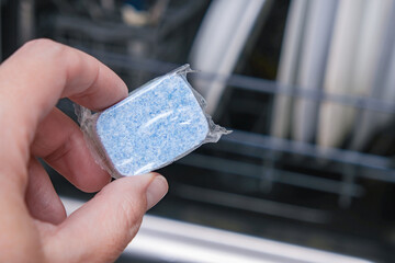 A hand holding blue dishwasher soap tablet in a water-soluble packaging on a dishashind machine background