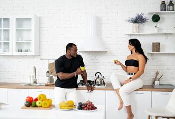 Dieting and healthy food concept. Beautiful young woman sitting near multiracial man in the kitchen with apple at the hands and chatting