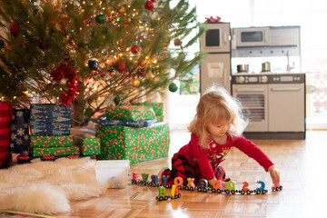 Little cute blonde toddler girl in pajamas playing with toys under Christmas tree on a Christmas...