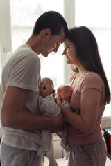 Loving family with newborn twins. Yong parents standing face to face near the window. Cute and tender pfoto of twins sleeping in mother's and father's arms. Siluette photo. Selective focus. Film grain
