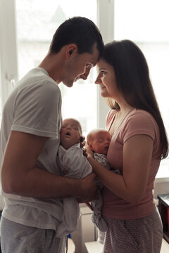 Loving family with newborn twins. Yong parents standing face to face near the window. Cute and tender pfoto of twins sleeping in mother's and father's arms. Siluette photo. Selective focus. Film grain