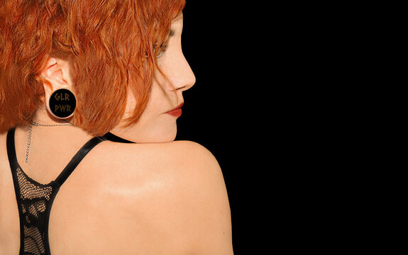 Back view of a sexy redhead woman looking over her shoulder with a GLR PWR spacer in her stretched ear. Beautiful model looking back wearing black bra and red lipstick. Isolated on  black