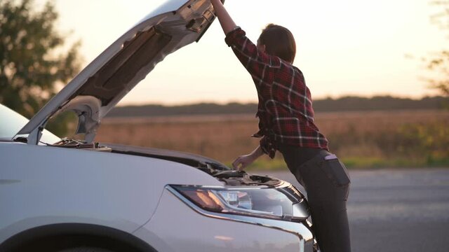 Car breakdown on road. Young attractive woman opens hood of car. Frustrated driver doesn't know what to do. Side view.