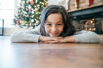 Portrait of happy girl laying on floor in front of Christmas tree resting chin on hands