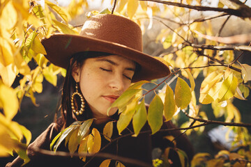 Portrait of stylish woman in hat and brown clothes posing among autumn leaves in warm sunny light. Fashionable young female standing at autumn tree branches in evening park or countryside