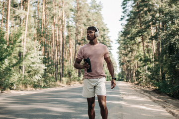 Sporty tired African American man runner, drinking water resting after training, on a city road in nature