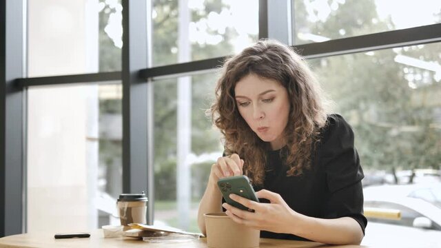 Curly haired young woman smiles looking into smartphone and eats from paper bowl sitting at wooden table in spacious light cafe