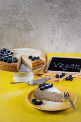 A vegan cheesecake with blueberries, a piece of cake on a plate and a chalk board with the words...