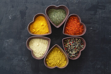 Different kind of spices on a black stone. Oriental spices in in heart shaped bowl, red peppers, curry powder, cinnamon powder, mint powder, colorful peppers. Flat lay, top view.