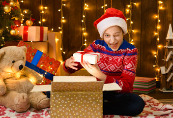 Fototapeta na wymiar Child girl posing in new year or christmas decoration, wearing a red sweater and a Santa hat, she opening a gift box and having fun. Holiday lights and lots of gifts, Christmas tree with toys.