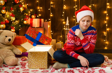 Fototapeta na wymiar Child girl posing in new year or christmas decoration, wearing a red sweater and a Santa hat, she opens a gift box. She is disappointed and has bad emotions.