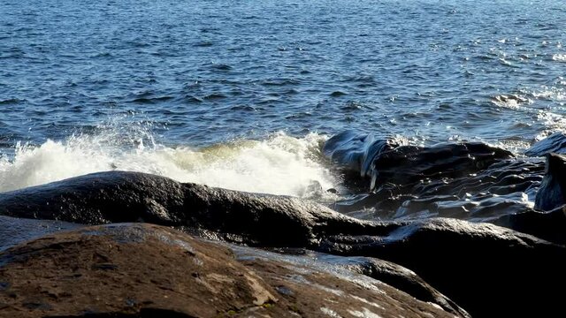 Waves breaking on the rocky north shore of Lake Superior in Minnesota on a beautiful sunny day, with white spray splashing on the rocks from the blue water. Slow motion clip with zoom out.