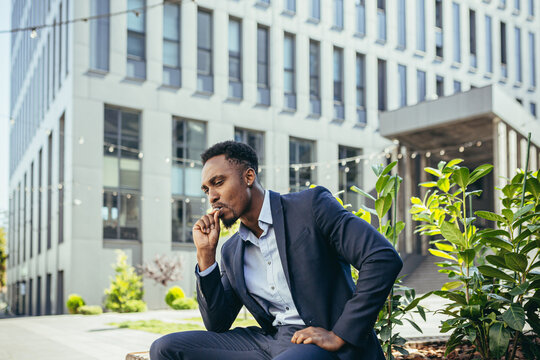 african american business man smoking cannabis outdoors sitting on city park bench on urban street background. Male employee Businessman. Office worker in suit Relieves stress a marijuana outside