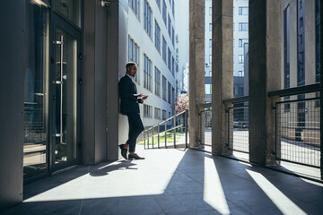 Fototapeta premium Male businessman near office reads news from tablet computer, african american freelancer in business suit near office outside
