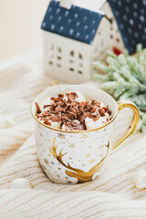 Hot chocolate cup with marshmallows on woolen plaid with Christmas decorations and small ceramic houses. Christmas beverage.