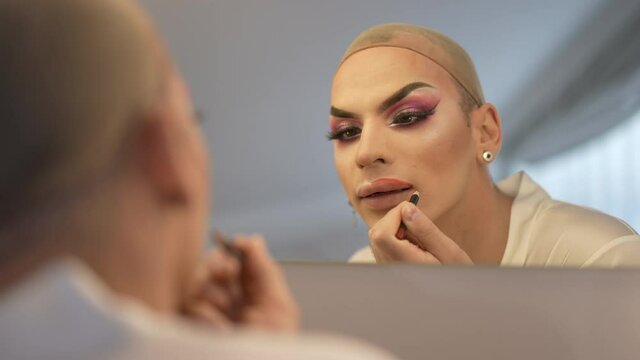 Reflection in mirror focused LGBT gender-fluid woman painting with lip pencil in slow motion. Young Caucasian queer person in wig cap doing makeup indoors. Individuality and lifestyle