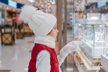 Boys in white fluffy hat looks into the store through the window and points something ih showcase in market.