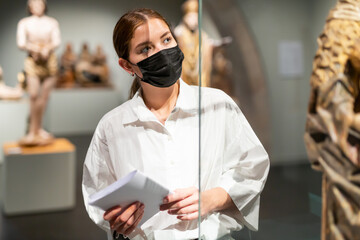 Portrait of young attractive woman in face mask visiting exhibit at sculpture hall in historical...