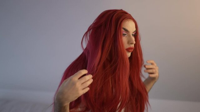Side view portrait focused transgender woman adjusting red wig standing indoors in white bathrobe. Concentrated proud Caucasian non-binary LGBT person getting ready to go out. Slow motion