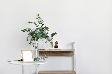 Vase with green eucalyptus branches and candle on shelf near white wall