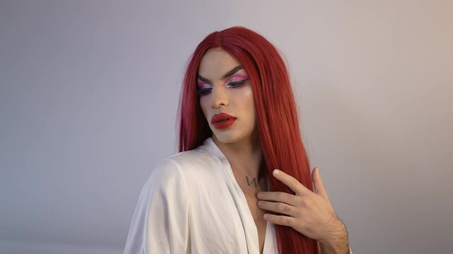 Front view portrait confident proud extravagant transgender woman brushing red long hair in slow motion. Portrait of Caucasian non-binary LGBT person in wig posing indoors at grey background