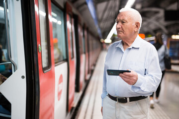 Elderly Caucasian man standing in metro station beside arrived train and holding smartphone in hand.