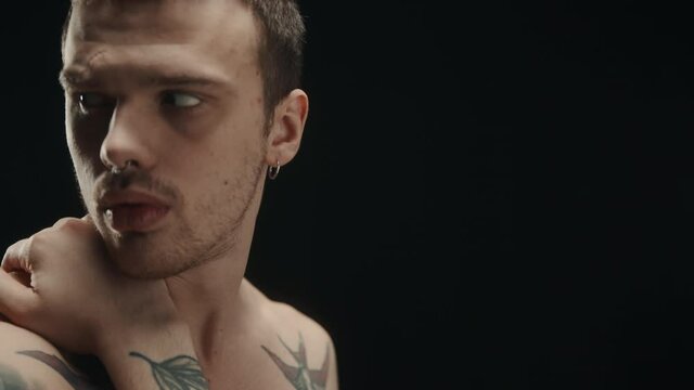Fashion portrait of brutal naked man in tattoos, having earring in his nose and ears, rubbing his hand over his neck, strong muscle guy posing in dark studio, looking in camera. 