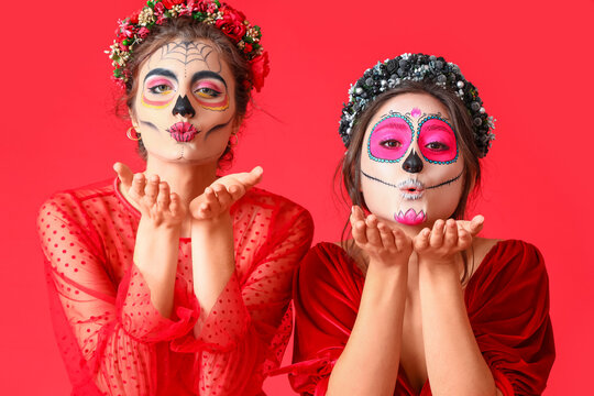Stylish women with painted skull on faces blowing kiss against color background. Celebration of Mexico's Day of the Dead (El Dia de Muertos)