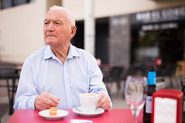 Fototapeta na wymiar European old man sitting at table in outdoor cafe with cup of coffee and cake.
