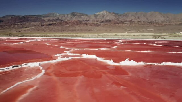 Aerial view of ancient dry salt lake in Death valley park, California USA. Pink salt lake marine, red water algae bloom with white peaks of natural salt and mountains landscape on motion background 4K