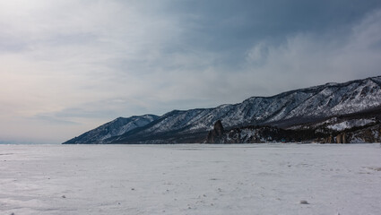A wooded mountain range against a blue sky. A bizarre rock devoid of vegetation is visible on a frozen and snow-covered lake. Baikal