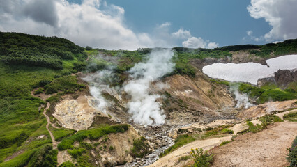Smoke from fumaroles rises above the mountain slopes. Paths and areas of melted snow are visible. A stream from a hot spring flows along a rocky bed. Blue sky. Kamchatka