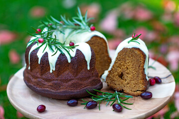 Christmas caramel cake, cutaway, decorated with white chocolate, cranberries and rosemary on a wooden stand among autumn leaves. Festive baked goods. Soft selective focus. Horizontal, close-up.