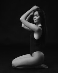Studio photo of pretty brunette woman wearing in a black bodysuit sitting on the studio floor on black background. Black and white