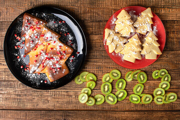 Christmas edible do-it-yourself baked Christmas trees made of pastry dough. Red pomegranate grain decoration, icing sugar like snow. New year 2022 is laid out from kiwi on wooden background, close up