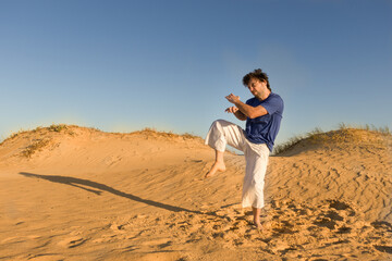Naklejka na ściany i meble joven practicando artes marciales al atardecer sobre dunas de arena, saltando 