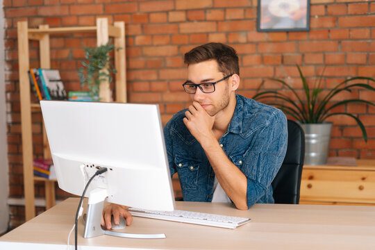 Medium Shot Of Handsome Young Man Wearing Stylish Glasses Working On Desktop Computer Sitting At Desk At Home Office In Room With Modern Interior. Freelance Programmer Working From Apartment.