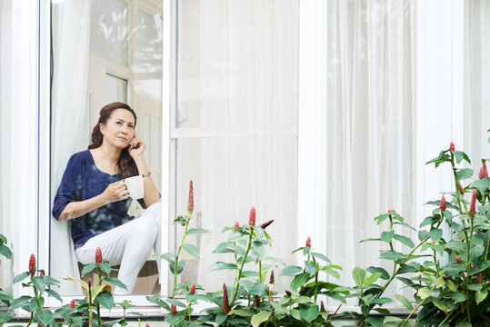 Pretty Serious Mature Woman Looking Through Apartment Window, Drinking Morning Coffee And Talking On Phone