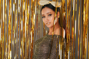 Portrait of attractive pretty Asian woman with bow on head standing at wall decorated with shiny golden tinsel and looking at camera
