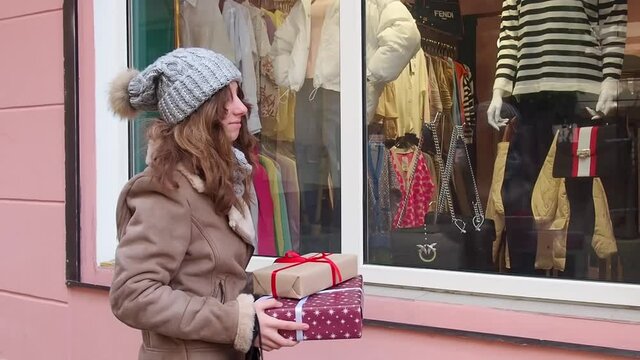 Teengirl in grey hat, scarf and brown jacket holding a set of christmas gifts near shops window. Happy young woman preparing holidays presents. Concept of shopping, giving and receiving gifts.