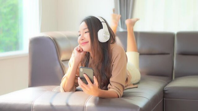 A young woman playfully kicks her feet while lying on the couch as she listens to music through her smartphone and headphones.