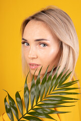 Naklejka premium Beauty portrait of a young, beautiful blonde woman with good skin in yellow clothes holding a green leaf on a yellow background