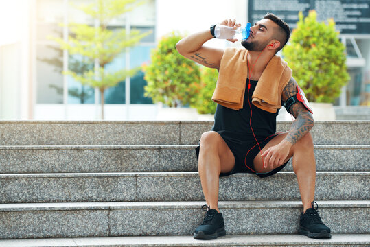 Fit handsome young man sitting on steps and drinking water after training outdoors
