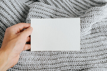 Close-up of hand showing white postcard mockup with blank space for text or bernding. Card template on background of knitted light scarf