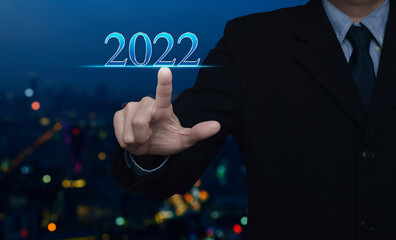 Businessman pressing 2022 text over blur colorful night light modern city tower and skyscraper, Happy new year 2022 calendar cover concept